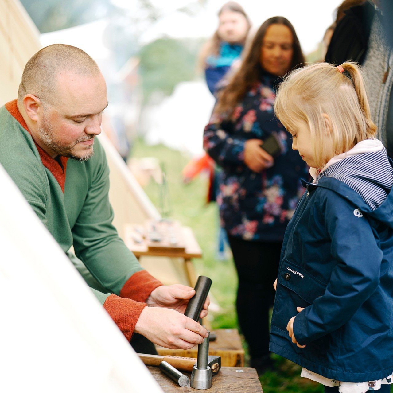 A person in early medieval-style clothing sits beside a coin‑striking tool and speaks with a child standing nearby, with other visitors and tents visible in the background.