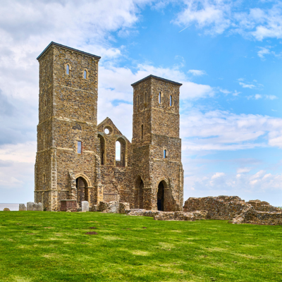 Reculver Towers, two stone towers of a 12th-century church, stand prominently on a grassy site overlooking the sea in Reculver, Kent. Scattered ruins surround the towers, and the sky above is partly cloudy with patches of blue.