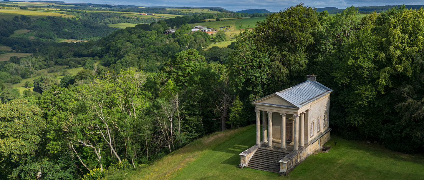 A small stone temple with columns at the front surrounded by green countryside
