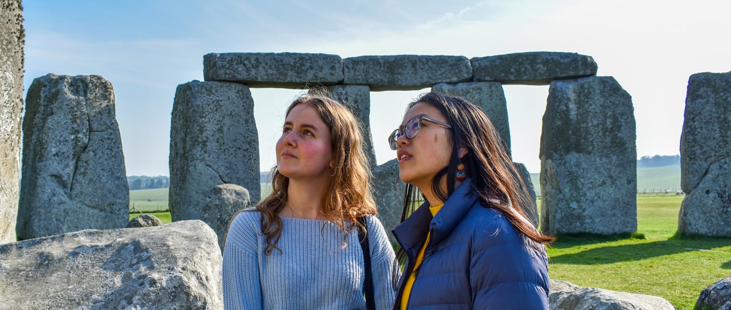 Two young women look up at Stonehenge on a sunny day, with the stone circle behind them.