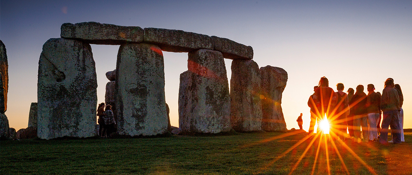 A group of people stand in front or Stonehenge as the sun crests the horizon
