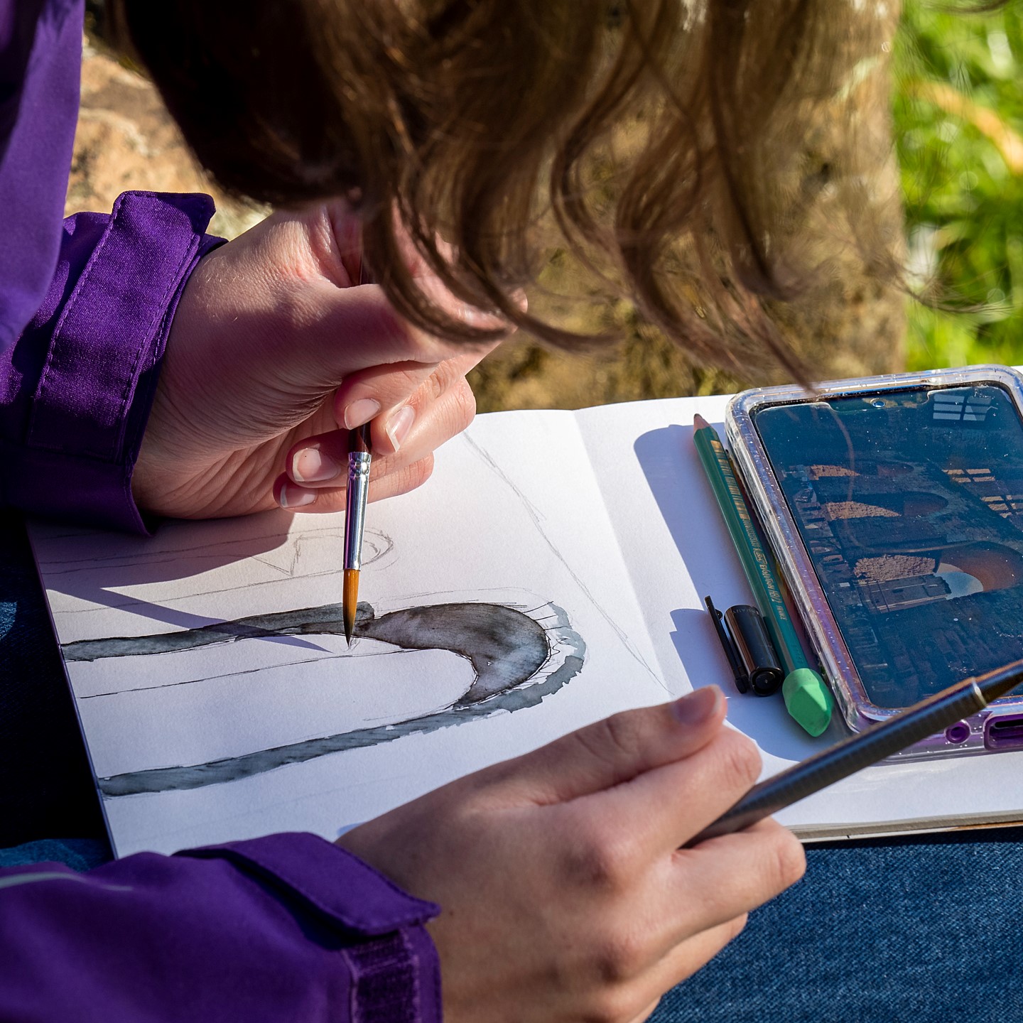 An artist paints a watercolour picture of a historic building whilst seated outside on grass.