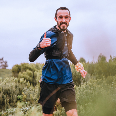 A person running on a trail through tall grass and bushes, wearing a blue shirt and black shorts, giving a thumbs-up.