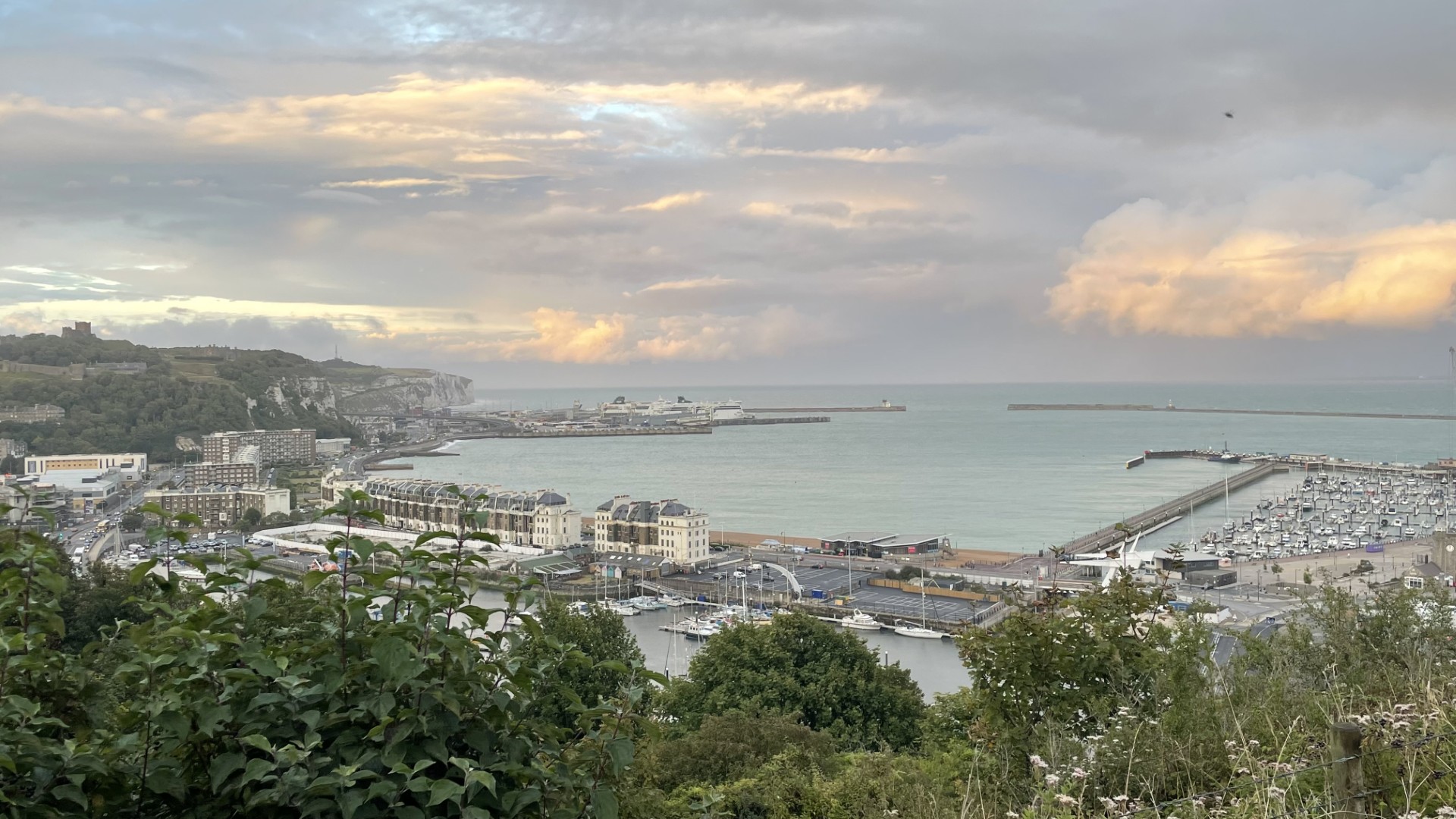 Panoramic view from Western Heights showing Dover town, the harbor, and Dover Castle. The scene includes a mix of urban buildings, coastal waters, and the historic castle perched prominently on a hill, all under a cloudy sky with patches of sunlight.