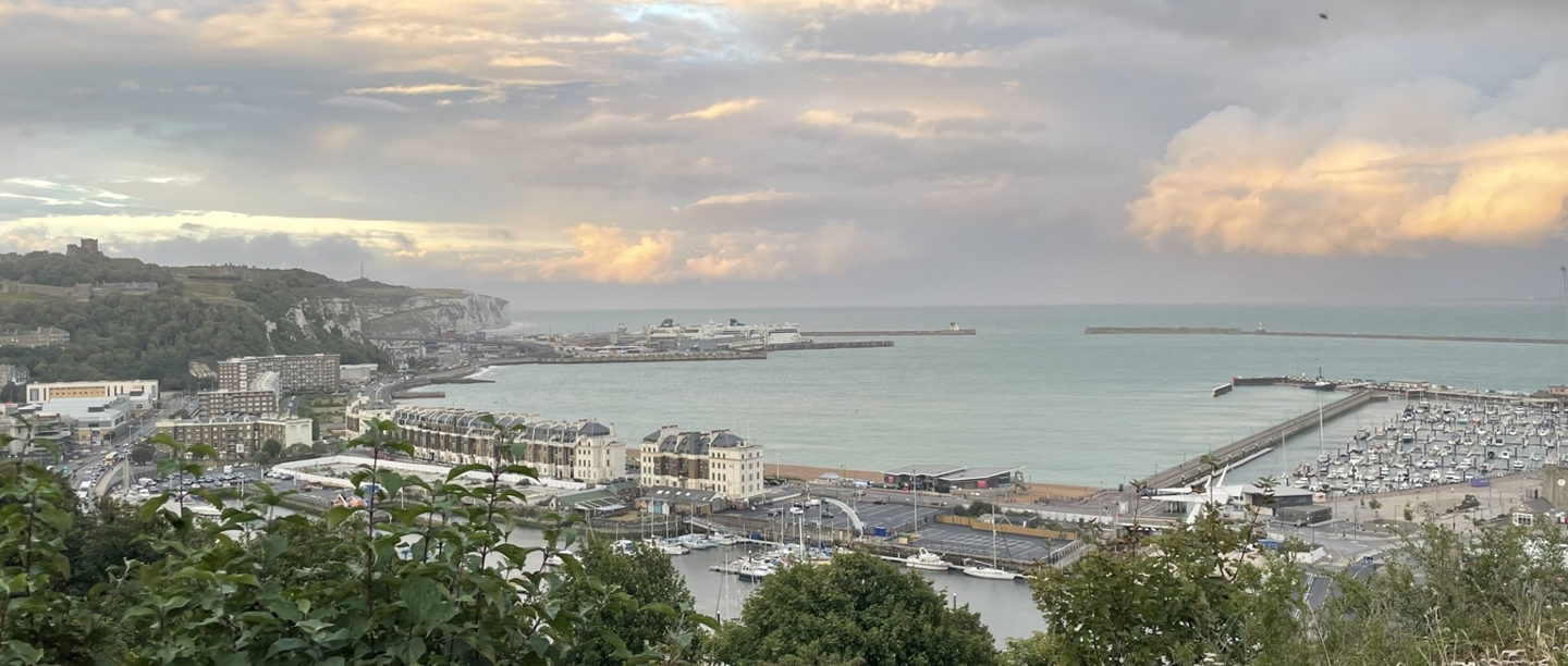 Panoramic view from Western Heights showing Dover town, the harbor, and Dover Castle. The scene includes a mix of urban buildings, coastal waters, and the historic castle perched prominently on a hill, all under a cloudy sky with patches of sunlight.