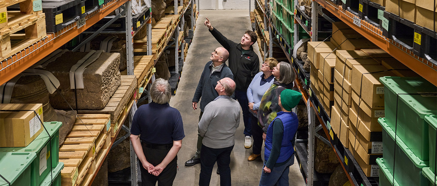 A man stands in front of a group of people in a storeroom pointing at one of the shelves