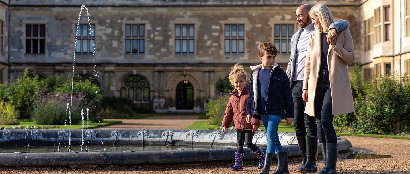 Two children, a man and a woman wearing coats and wellies walk in front of a large country house (Audley End House)