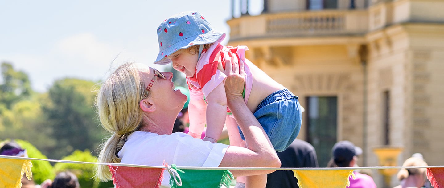 A woman holds a baby up smiling next to a string of colourful bunting with Osborne in the background.