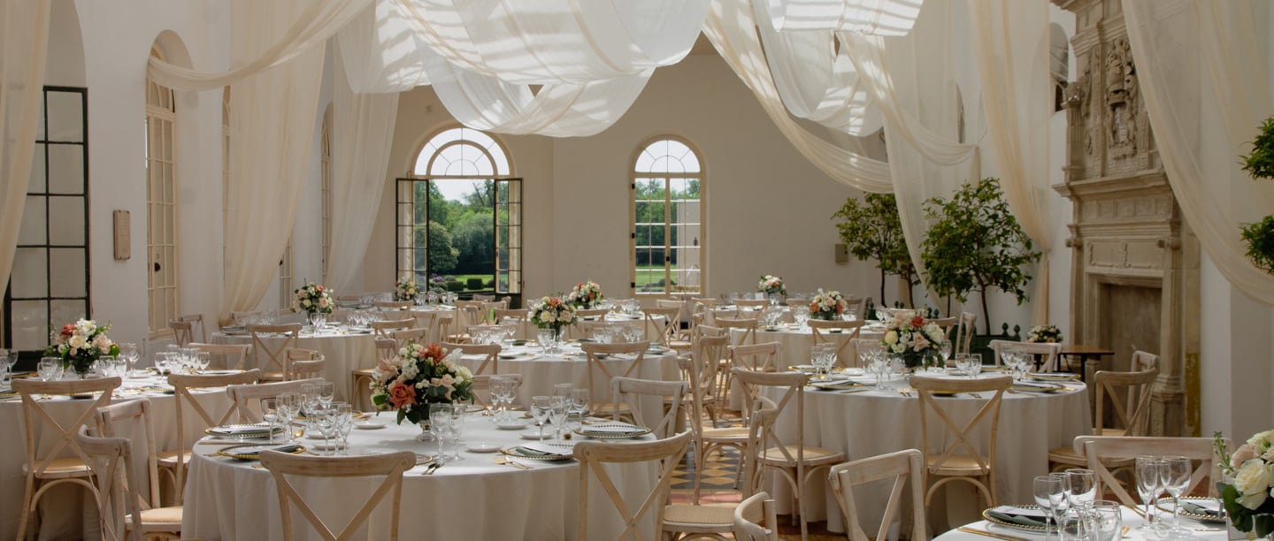 A wedding venue set up with white draped fabric hanging from the ceiling, large circular tables with floral displays on with a view of the gardens outside.