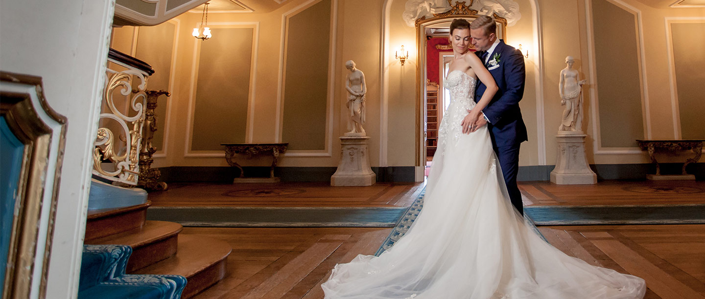 A man in a suit and woman in white wedding dress embrace in a grand hallway
