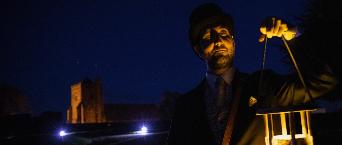 Image: Photo of a person dressed in Victorian costume holding a lantern in the dark