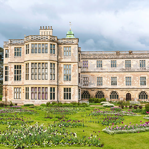 A grand mansion with three storeys beyond a green lawn and colourful flowers