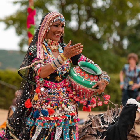 A woman in colourful traditional clothing dances outdoors, holding a decorated pot. She is smiling and wearing vibrant jewellery.