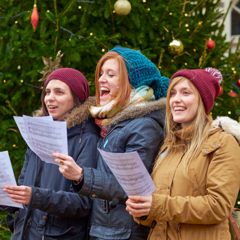Three young women singing Christmas carols outdoors in front of a decorated Christmas tree. They are dressed warmly in coats, scarves, and hats, holding sheet music and smiling as they sing.