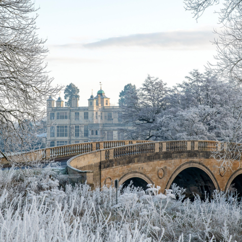 A stone bridge curves over a frosty landscape, with trees and bushes covered in white frost. In the background, the turrets of Audley End house can be seen behind the trees.