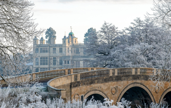A stone bridge curves over a frosty landscape, with trees and bushes covered in white frost. In the background, the turrets of Audley End house can be seen behind the trees. A stone bridge curves over a frosty landscape, with trees and bushes covered in white frost. In the background, the turrets of Audley End house can be seen behind the trees.