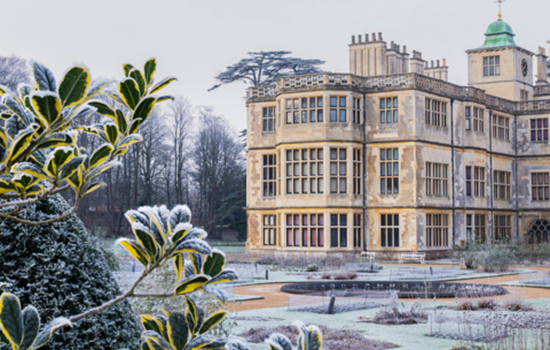 A grand historic mansion with tall chimneys and large windows, surrounded by formal gardens lightly dusted with frost. The foreground shows frosted evergreen leaves, and bare trees stand in the background under an overcast winter sky A grand historic mansion with tall chimneys and large windows, surrounded by formal gardens lightly dusted with frost. The foreground shows frosted evergreen leaves, and bare trees stand in the background under an overcast winter sky