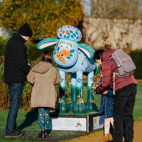 A group of adults look at a large light blue and orange sculpture of Shaun the Sheep in the grounds of Wrest Park.