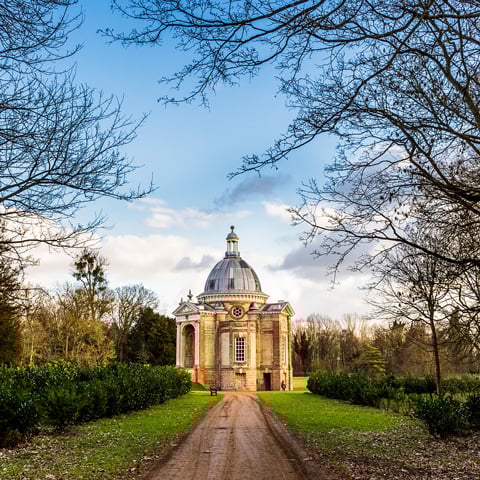 A view of the gardens at Wrest Park underneath blue sky with a circular dome roof building in the foreground. 