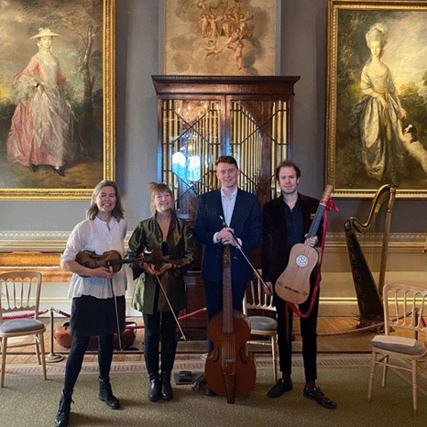 Four people holding string instruments smile for the camera. They are stood in a large room at Kenwood House and are stood infront of two large portraits of women.