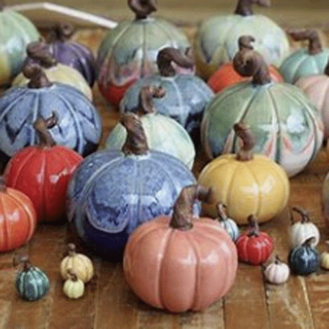 A collection of decorative clay pumpkins in various colors and patterns, including shades of blue, green, yellow, red, and pink, arranged on a wooden surface. Each pumpkin has a brown stem and they vary in size.