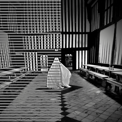A black and white image of a figure covered in a large white sheet in a large striped timber-framed hall. 