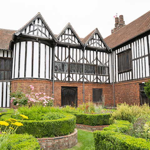 A black and white timber framed hall overlooking a cortyard filled with small hedges and flowers.