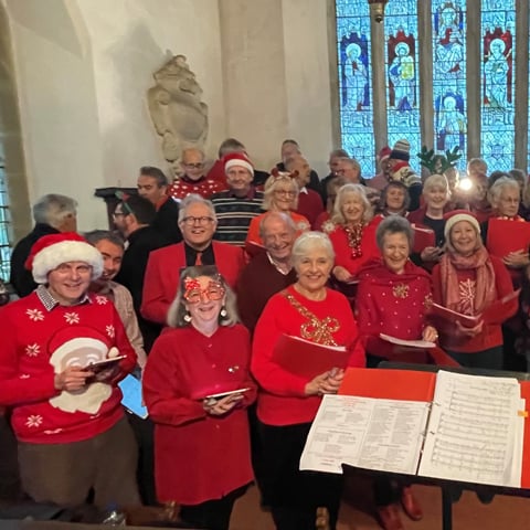The Roaring Megs choir stand smiling in a Chapel wearing red Christmas jumpers.