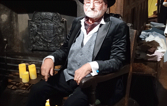 A man dressed in a grey waistcoat, black suit and a black top hat sits on a wooden chair next to lit candles. A man dressed in a grey waistcoat, black suit and a black top hat sits on a wooden chair next to lit candles.
