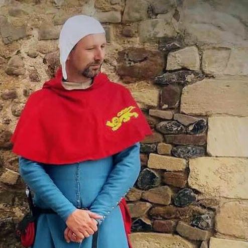 A person dressed in a medieval historical blue and red costume stands with their hands together in front of a stone wall.