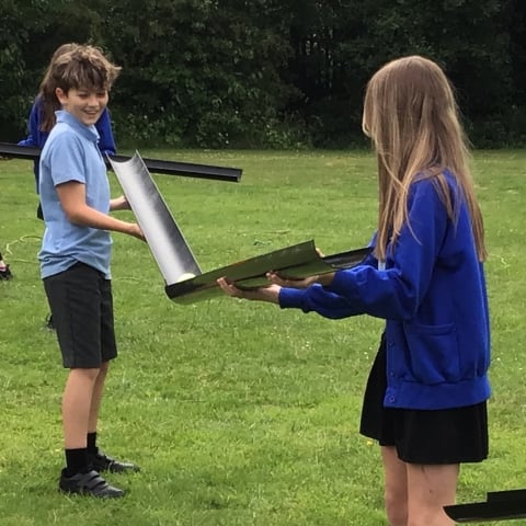 Two older children stand on a field, smiling, holding plastic guttering to pass a tennis ball between them. They both wear blue school uniform.