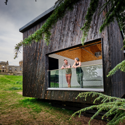 Two people standing on the balcony of a modern wooden structure with a large open window, set in a grassy area near Witley Court, visible in the background