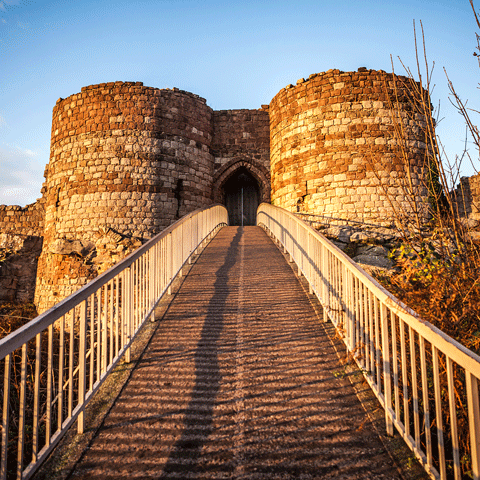 Looking towards a castle bridge entrance perched on a hill, set against a clear blue sky in the background.