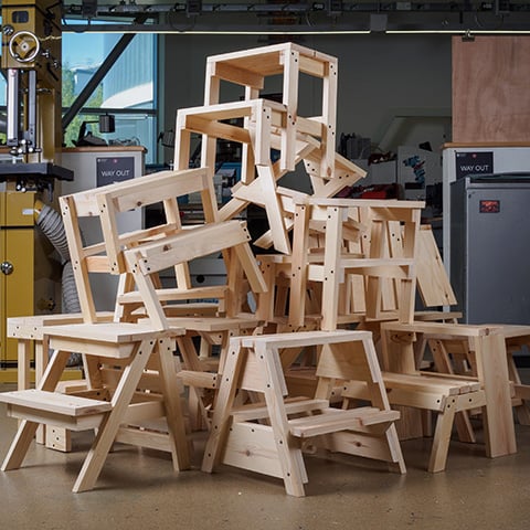 A large stack of unfinished wooden step stools piled on top of one another in a workshop with various tools and equipment visible in the background.