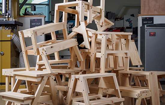 A large stack of unfinished wooden step stools piled on top of one another in a workshop with various tools and equipment visible in the background. A large stack of unfinished wooden step stools piled on top of one another in a workshop with various tools and equipment visible in the background.