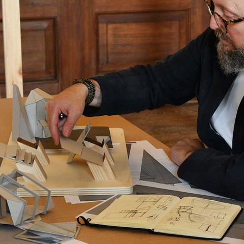 A man with glasses holds a cardboard and wooden abstract model on a wooden desk filled with papers and drawings.