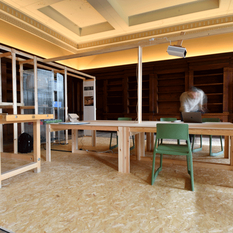 A workshop with wooden tables and green chairs. There is a blurred out person stood over one of the tables.