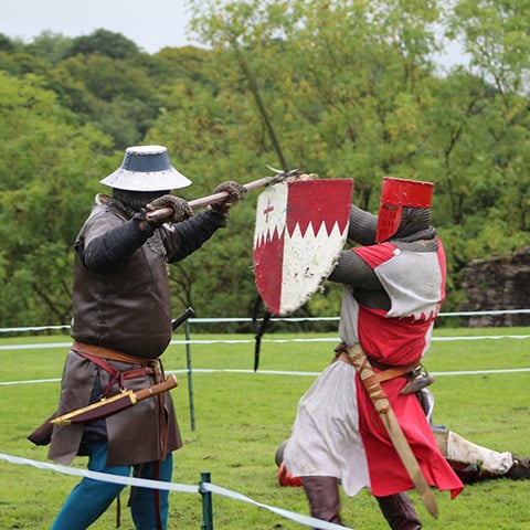 Two people engaged in battle combat re-enactment. They are dressed in historical armour and clothing, wielding swords and shields.