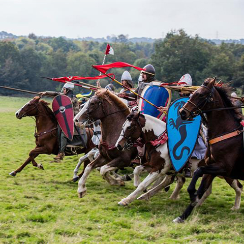 A group of reenactors ride horses, wearing battle armour and holding shields.
