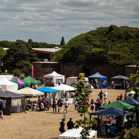 Outdoor craft fair with rows of colourful canopy stalls and visitors in a gravel courtyard beside a red-brick building and trees under a cloudy sky.