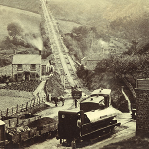A sepia photograph of a rural railway scene with steam trains, coal wagons, and a steep incline track leading up a hillside, surrounded by stone buildings and fields.
