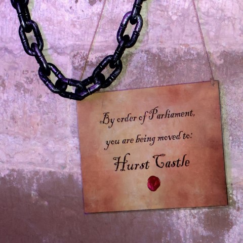 A dark iron chain hangs next to a card against a bleak stone background. The card reads - By order of Parliament, you are being moved to: Hurst Castle. A dark iron chain hangs next to a card against a bleak stone background. The card reads - By order of Parliament, you are being moved to: Hurst Castle.