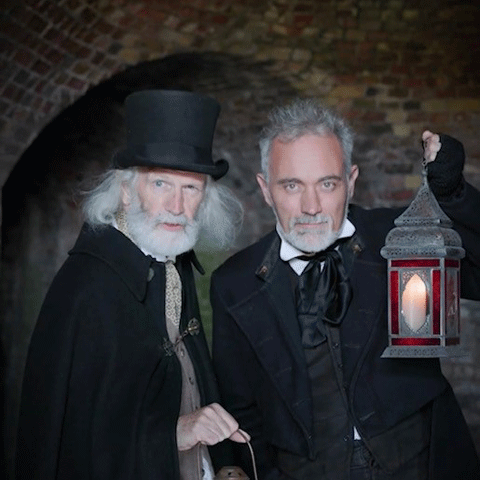 Two men dressed in Victorian style historical costumes holding lanterns in a dark setting.