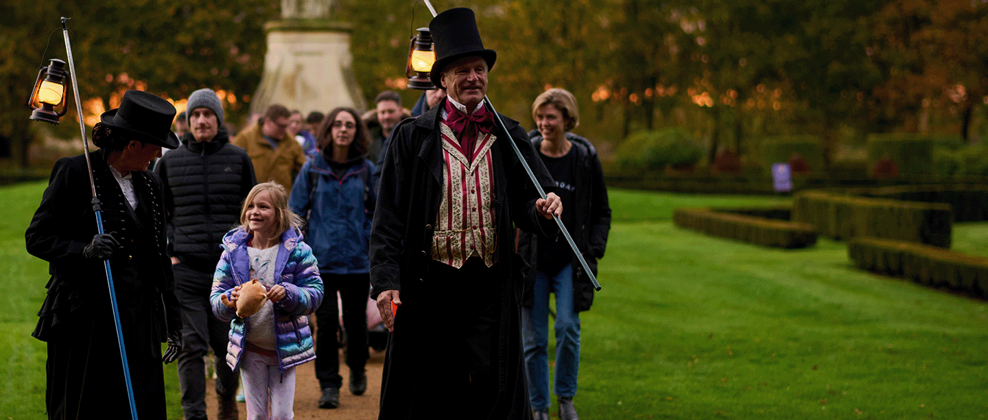 Two people in period clothing carrying lanterns in a garden lead a group of adults and children at dusk.