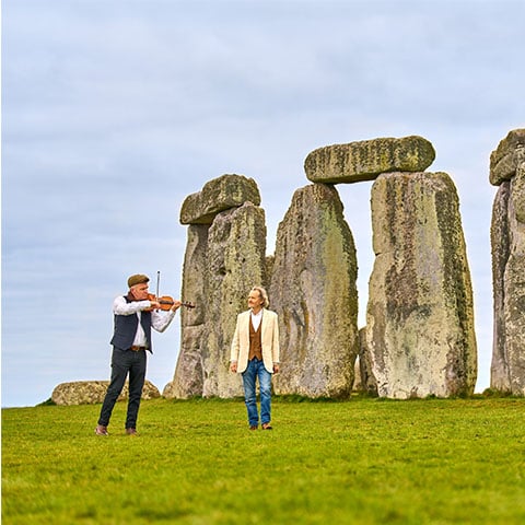 Anton Lesser walking in front of Stonehenge with a musician playing the violin