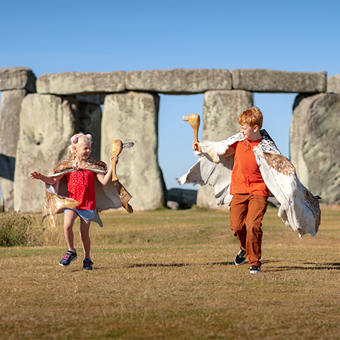 Two children run in front of Stonehenge dressed in fake furs and carrying pre-historic-style clubs