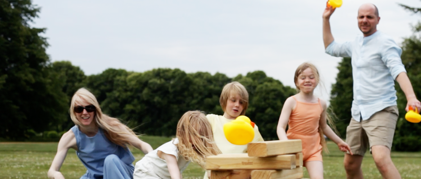 A family of three children and two adults play giant Jenga outdoors, playfully throwing rubber ducks and having fun in a large field.