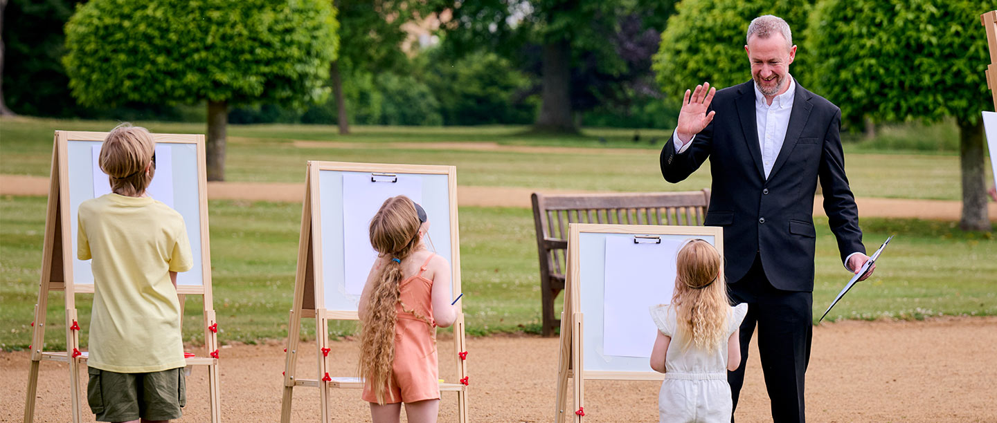 Three children stand at easels while a man in a suit (Taskmaster's Alex Horne) waves at them