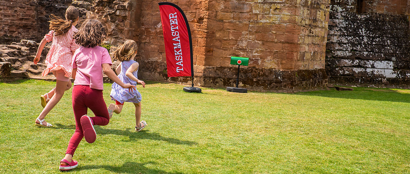 Three girls run towards a Taskmaster banner flag