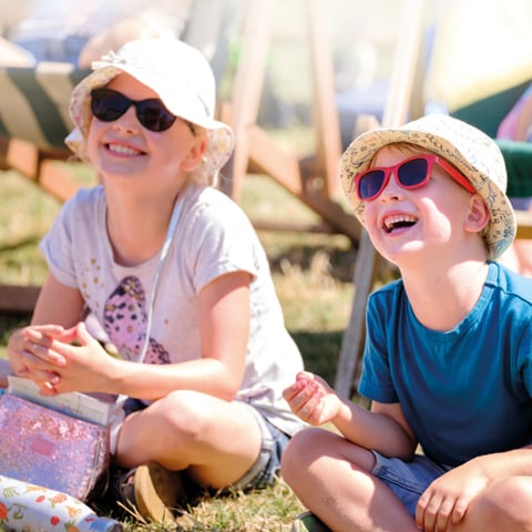 Two children wearing sunglasses and sun hates smile as they sit on the grass with wooden deckchairs in the background. 